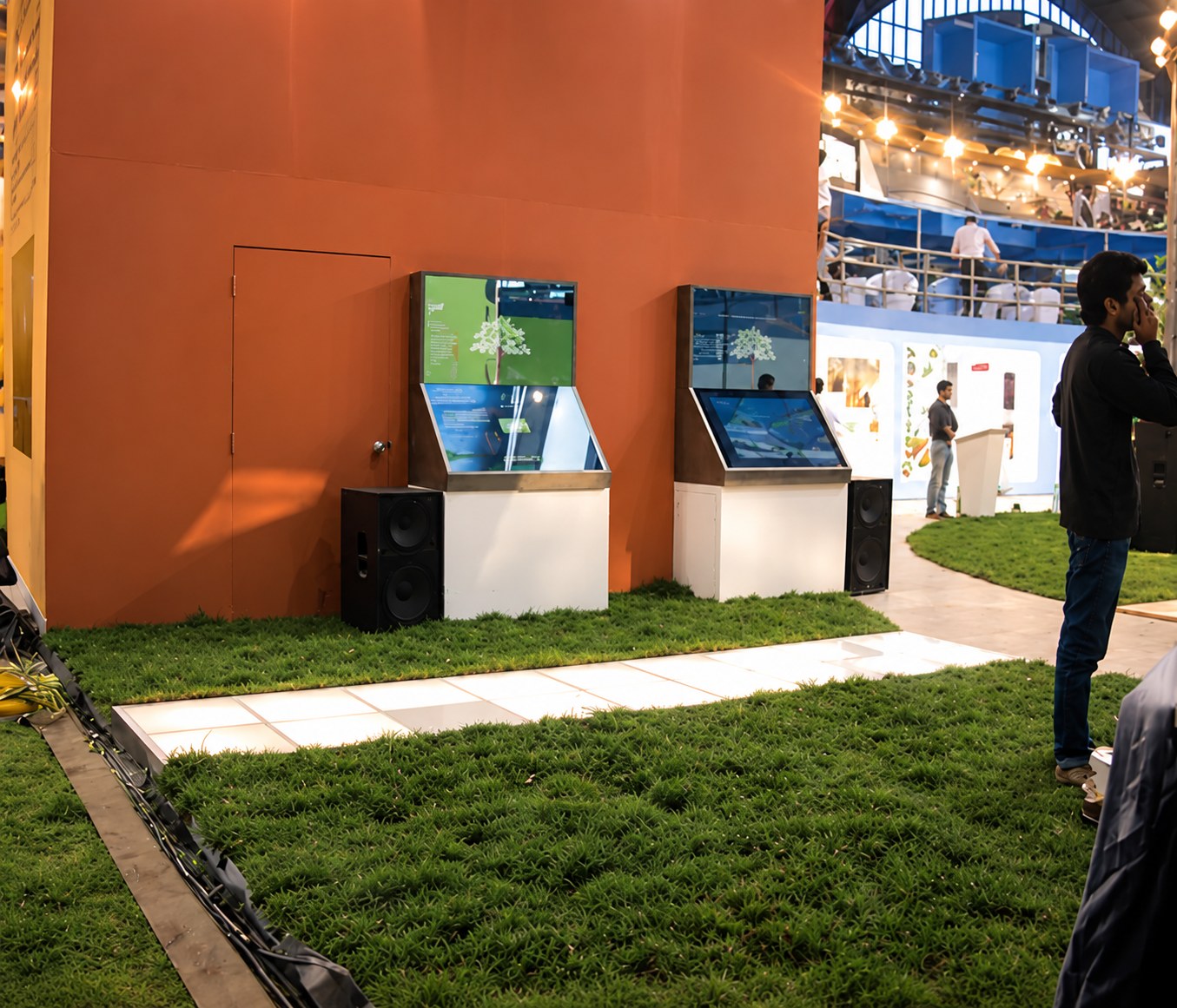 Two touchscreen game kiosks against an orange wall at an exhibition, on a lawn of artificial grass.