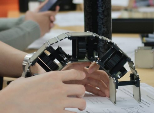 Hands bending the toy model of the wall into an arch on a table.