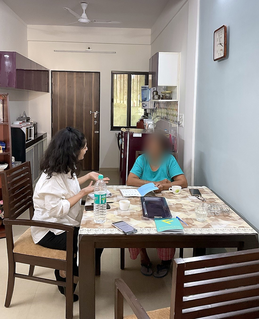 An interview at a dining table at home: the researcher talks with an older woman who holds her open booklet.