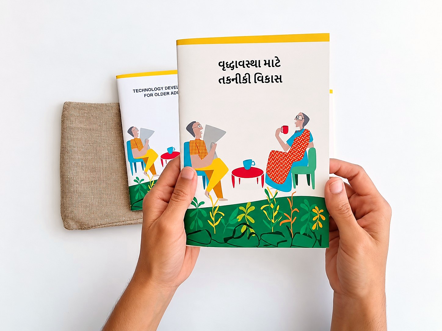 Hands holding the Gujarati edition of the booklet, its cover showing an older man reading a newspaper and an older woman in a sari with a cup of tea, beside the English edition and a jute pouch.